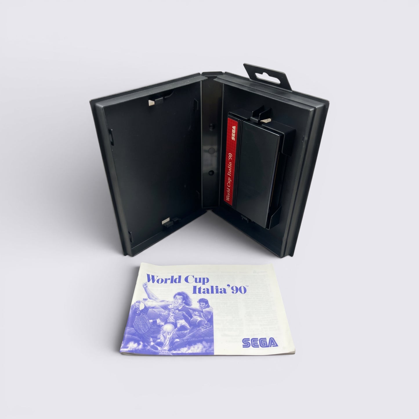 MrPixel World Cup Italia 90 Sega game cartridge in black case with original instruction manual on grey surface